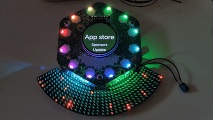 Looping video of two Matrix Hexpansions with the lite-loop daughter board plugged into the bottom expansion slots of a 2024 Tildagon Badge. The LED matrix displays a starfield pattern where specks of light move outwards from the badge center. Each hexpansion is a grid of 156 tiny orange LEDs on a black PCB arranged in a 9-row wide circular strip tangentially to the badge outline. The inner-most row has a gap of three LEDs on each side, the remaining rows join seamlessly with the adjacent board.
